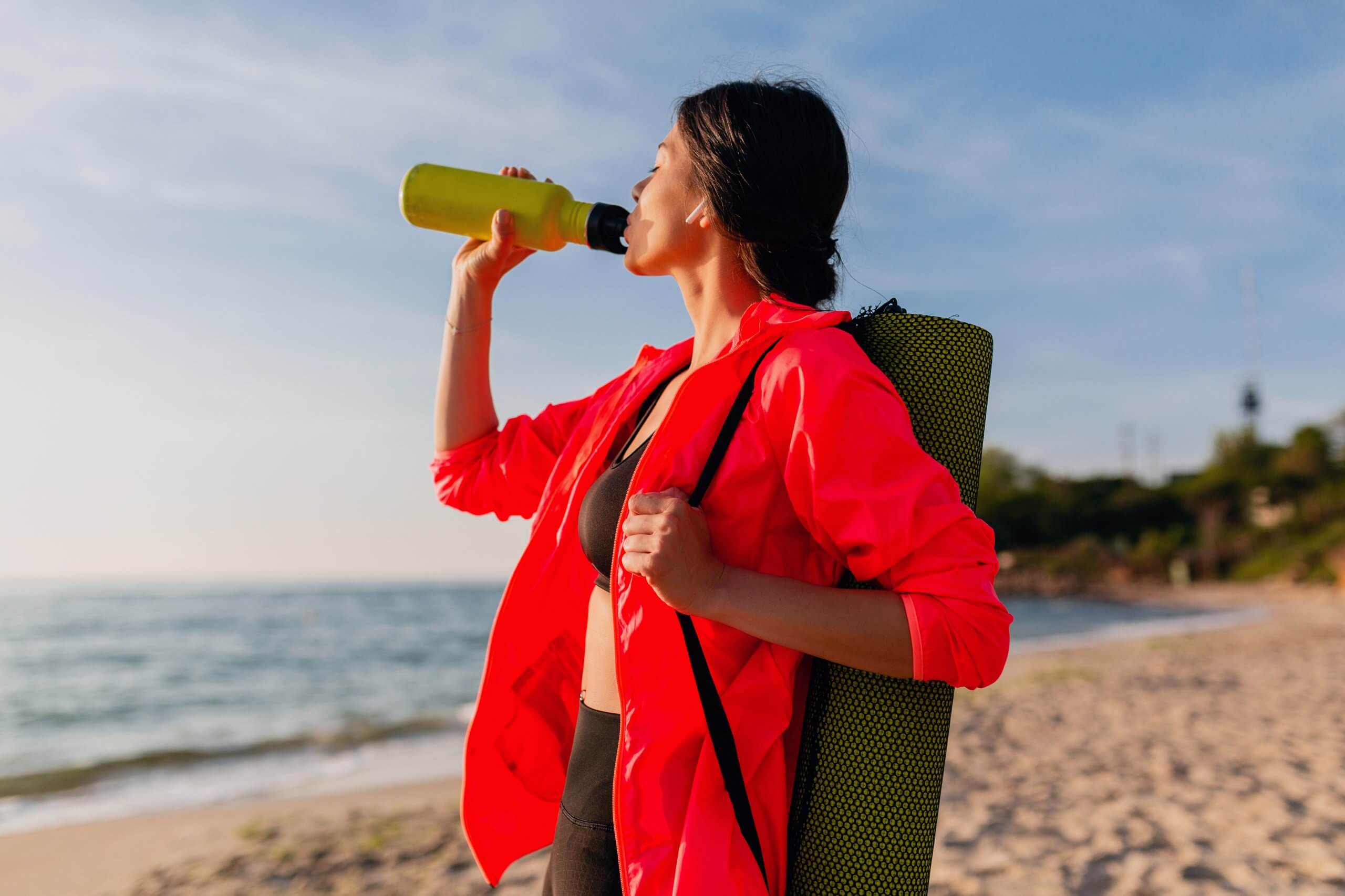 young attractive smiling woman doing sports in morning sunrise on sea beach holding yoga mat and bottle of water, healthy lifestyle, listening to music on earphones, wearing pink windbreaker jacket
